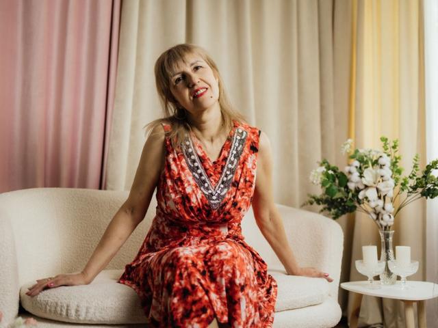 PattySanny, eine Frau mittleren Alters mit blonden Haaren, trägt ein rotes und schwarzes Kleid mit Muster und sitzt auf einem weißen Sofa, lächelnd, mit den Händen auf den Kissen. Auf dem Beistelltisch stehen Blumen und Kerzen..