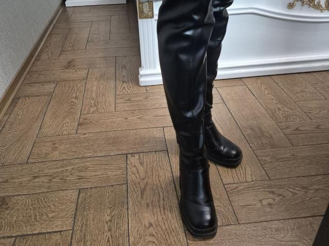 Les bottes en cuir noir et le pantalon d'Anshante sont posés sur un plancher en bois à motif chevrons. Les bottes sont brillantes et ont un petit talon. Le pantalon est légèrement froissé..