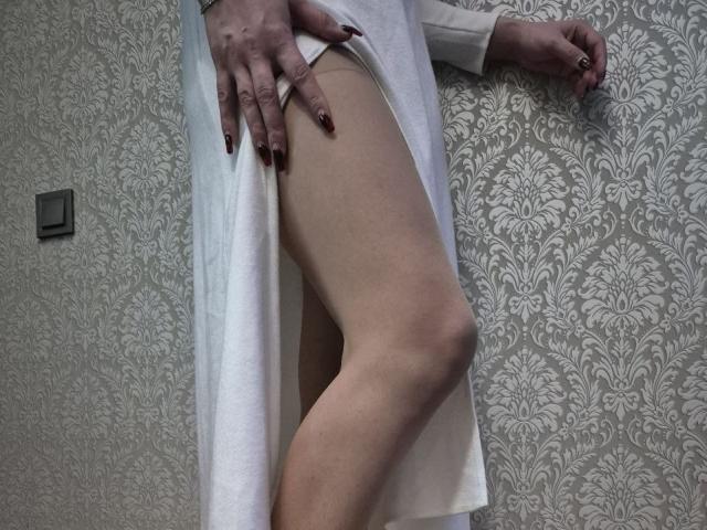 La robe blanche d'Anshante est remontée, exposant sa cuisse nue et ses fesses. Elle est debout, une main sur sa jambe, montrant des ongles d'un rouge foncé. Sa jambe est légèrement pliée..