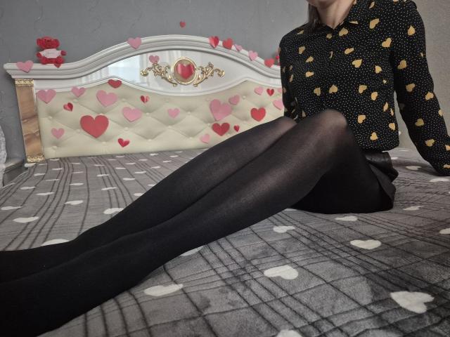 Anshante, portant une chemise noire à pois, avec des cœurs jaunes, des collants noirs et une jupe courte, assise sur un lit avec une couverture grise à motifs de cœurs, montrant ses jambes et ses fesses..