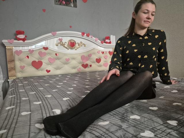 Anshante, avec ses seins couverts, portant une chemise noire à pois et des collants noirs, assise sur un lit à motifs de cœur, a l'air détendue avec les jambes tendues..
