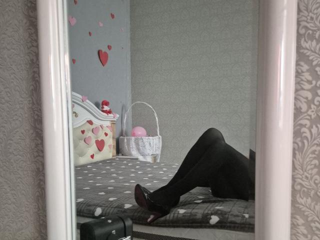 Anshante's black pantyhose-clad legs are spread on a gray polka-dot bed, showing her purple heels. A pink basket and heart-decorated white dresser are in the background.