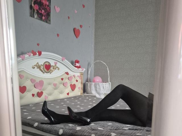 Las piernas de Anshante, cubiertas con medias negras, y sus zapatos negros brillantes están sobre una cama gris con lunares, con pegatinas de corazones rosas y una cesta blanca en una habitación gris..