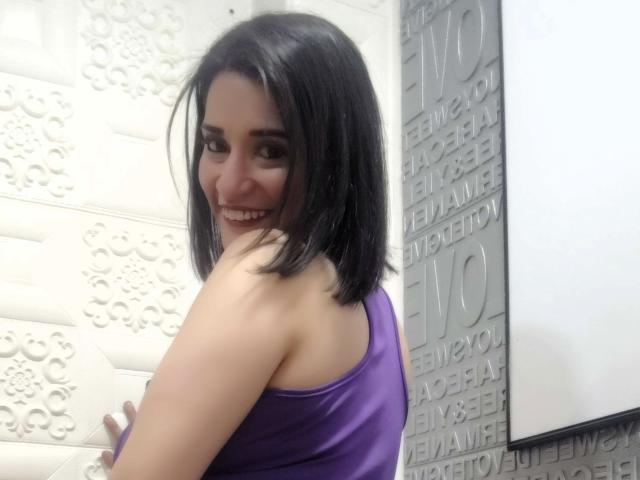 MaraRoberttss with shoulder-length black hair, smiling, wearing a purple satin tank top, showing her back and looking over her shoulder, standing against a white textured wall.