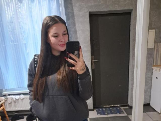 FabinaBae mit langen, braunen Haaren, die einen grauen Hoodie trägt, macht ein Selfie mit ihrem schwarzen Handy, lächelt und zeigt ihr Hinterteil in engen Jeans, in einem Zimmer..