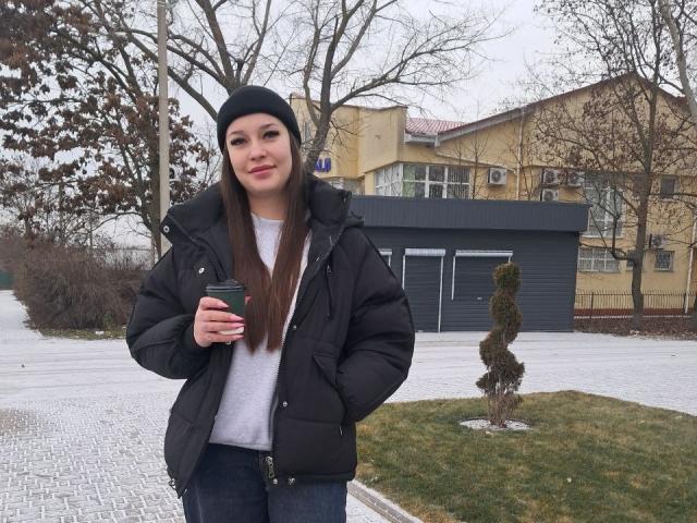 FabinaBae com cabelos castanhos longos, gorro preto, jaqueta preta acolchoada, camisa branca, jeans azuis, segurando uma caneca verde, em frente ao jardim, árvores sem folhas, arbusto pequeno, céu nublado..