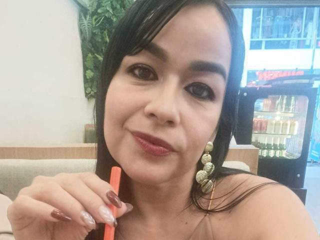 TamaraFoxy with dark hair, holding a red straw, wearing gold earrings, and pink lipstick, smiling at the camera, showing her painted nails, in a relaxed indoor setting.