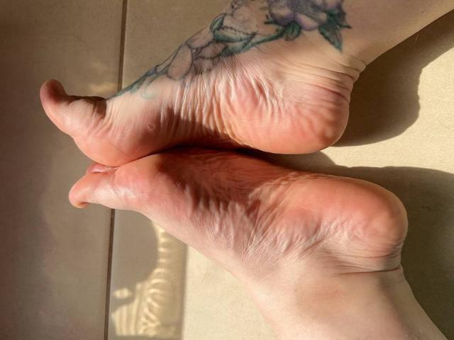 GoddessKisica's soles, showing wrinkled skin and a floral tattoo on her left foot, with sunlight highlighting her toes and the texture of her skin.