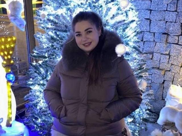 Melany Shaw mit dunkelbraunen Haaren, lächelnd, trägt einen dicken, braunen Mantel mit Fellbesatz, die Hände in den Taschen, und steht vor einem blau beleuchteten, schneebedeckten Weihnachtsbaum..