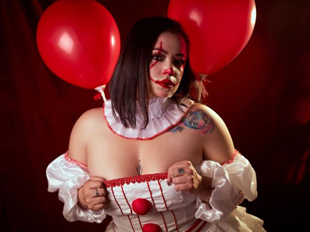SamanthaVenus con un palloncino rosso, un costume bianco con una spalla scoperta, e trucco rosso, che mostra il seno, tiene il costume, con tatuaggi visibili sulla spalla destra, capelli castano scuro, sfondo rosso..