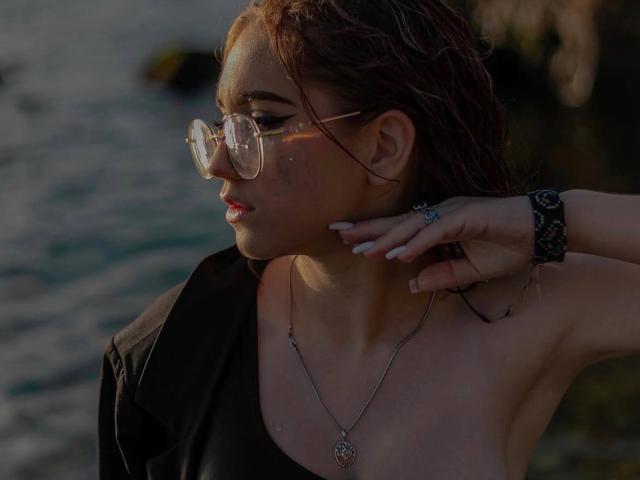 FellDivain with glasses, red hair, and white nails touches her neck, wearing a black top and silver necklace, showing her tits slightly. Wearing a black wristband, by the water.