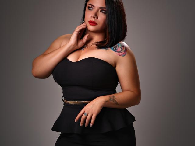 SamanthaVenus tiene senos grandes, un vestido negro sin tirantes y un tatuaje en el hombro derecho. Está tocándose la barbilla con lápiz labial rojo y lleva un cinturón marrón..