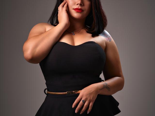 SamanthaVenus with black dress, red lips, hand on cheek, tattoo on right shoulder, tan belt, visible cleavage, medium breasts, dark hair, gray background.