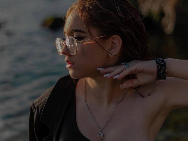 FellDivain with wet brown hair, wearing round glasses, black shirt, and necklace, has a ring on her finger and a black watch, by a dark, reflective water surface.