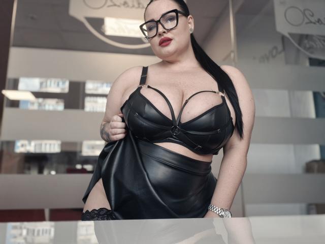 GeorgiaLerox, vestindo lingerie de couro preto, com seios grandes e um rabo de cavalo preto comprido, usando óculos, segurando uma saia de couro, em pé em uma sala moderna, mostrando uma tatuagem na mão..
