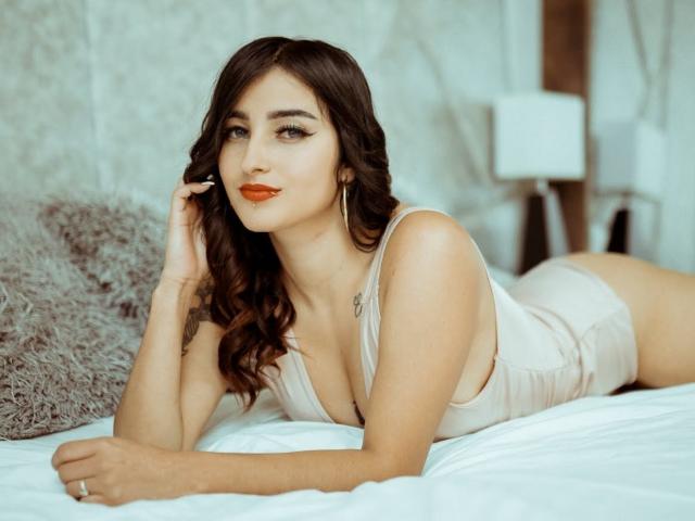 LisaNahyan is lying on a bed in a beige tank top and shorts, with wavy brown hair, red lipstick, and hoop earrings, showing her tits and a cute smile.