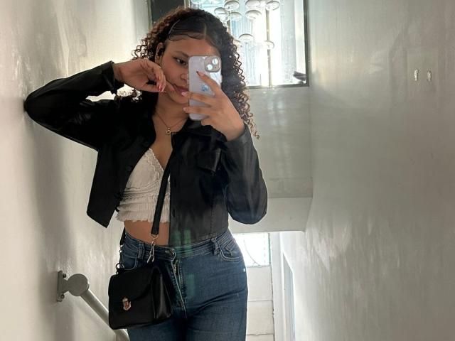 Sofia, com cabelo cacheado, jaqueta preta, blusa branca com renda, jeans azuis, bolsa preta, segurando o celular, selfie no espelho, mão perto do rosto, bumbum pequeno, seios médios, paredes brancas, escadas..