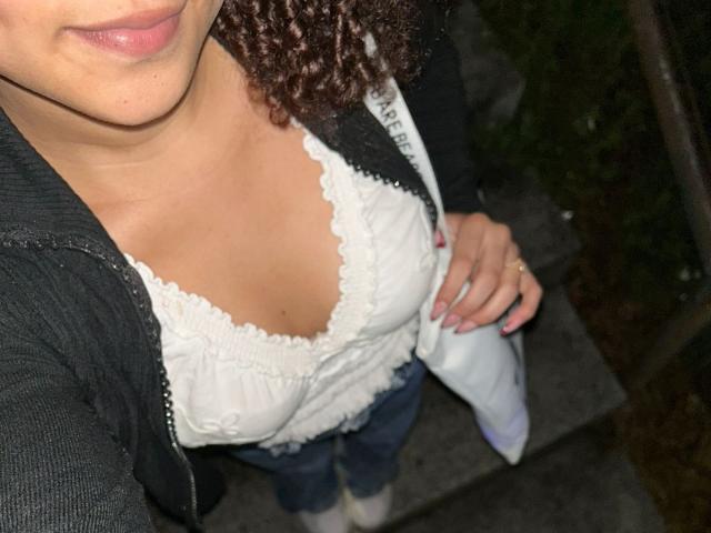 SofiaPassionne with curly brown hair, wearing a black jacket, white lace top, showing her boobs, blue jeans, and white socks, holding her right hand down. Night shot.