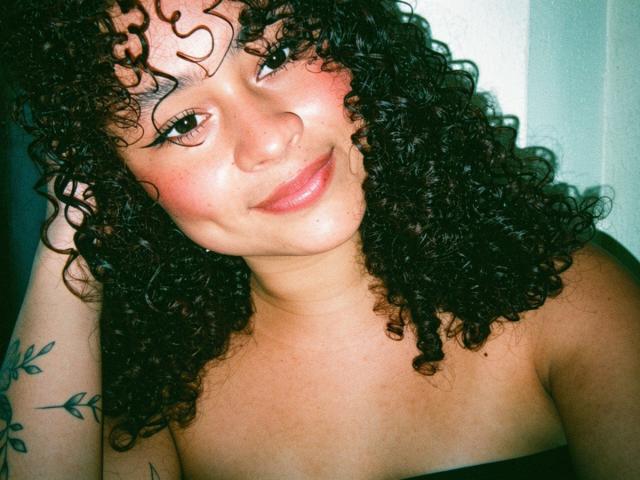 SofiaPassionne tem cabelos pretos encaracolados, pele morena e um sorriso discreto com lábios rosados. Ela está sem blusa, mostrando seus pequenos seios redondos e uma tatuagem no braço esquerdo..