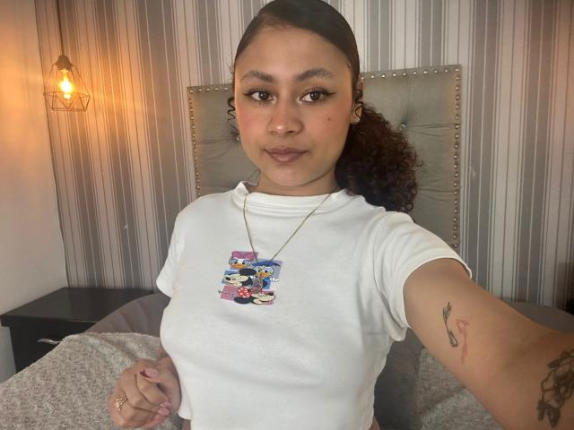 Sofia a les cheveux bruns bouclés, une peau marron clair, et porte un t-shirt blanc avec un collier en forme de voiture. Elle se touche les fesses et a un tatouage sur le bras..