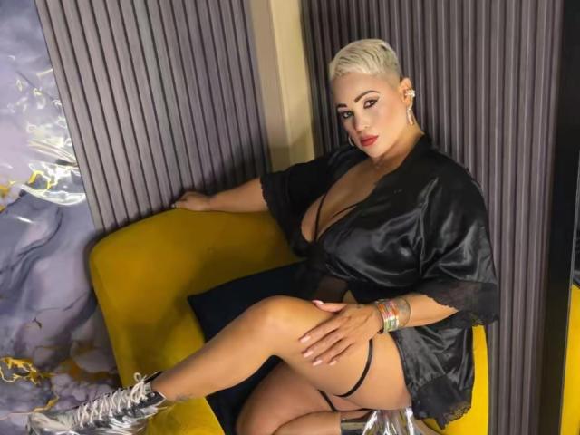 GianellaCox with short platinum blonde hair, wearing a black satin robe, showing big tits, sitting on a yellow chair, silver boots, black thong, and colorful bracelets.