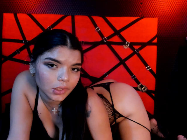 AbbyBlaze with long black hair, black lingerie, cross necklace, and a serious face, on all fours, showing her ass and a tattoo on her upper arm, in a red-lit room.