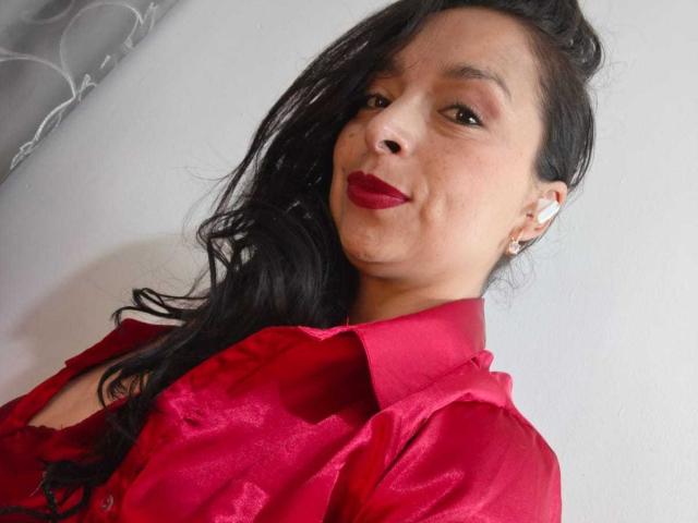 StangelarRivera, con cabello oscuro y ondulado, lápiz labial rojo y una camisa de satén roja, muestra una ligera sonrisa. Lleva aretes blancos y tiene un aro nasal discreto..