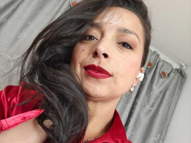 StangelarRivera, con capelli scuri e ondulati, rossetto rosso, lentiggini e orecchini bianchi, indossa una camicia rossa, guarda leggermente di lato con un sorriso appena accennato..