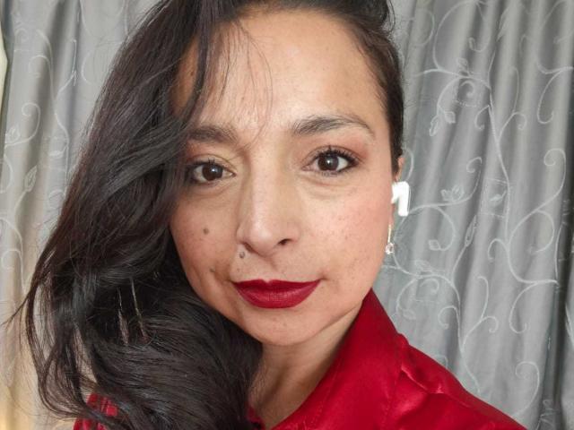 StangelarRivera has dark, wavy hair, red lipstick, and a small beauty mark on her left cheek. She's wearing a red shirt and white earrings. Her brown eyes are slightly upturned.