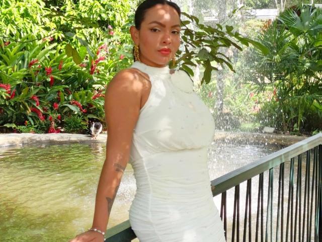 JessicaFiore with big tits in a white, sleeveless, ruched dress, standing by a water feature, wearing red lipstick, gold earrings, and a bracelet, in a sunny, green garden.