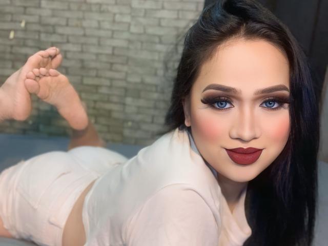 AmaraHugeCock liegt auf dem Bauch, trägt weiße Kleidung, zeigt ihr Gesäß, hat dunkles Make-up, roten Lippenstift und blaue Augen und schaut in die Kamera..