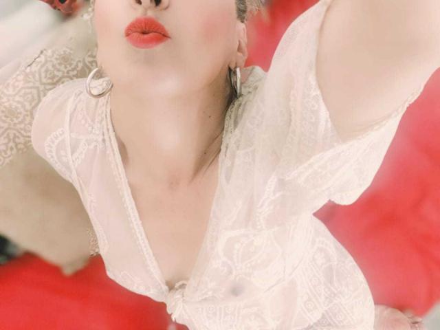 SammyDesire, con labios rojos, vistiendo una blusa blanca de encaje transparente que deja ver sus pequeños senos, con aros en las orejas y piel clara, tomando un selfie, con los brazos levantados, sobre una superficie roja..