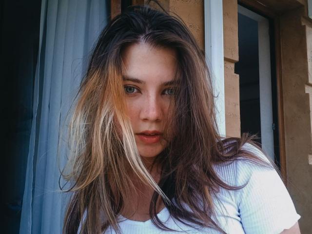 Mettis, con capelli biondo scuro e castani, occhi azzurri e una maglietta bianca, con un'espressione seria, in piedi vicino a una porta, con i capelli disordinati che le coprono parzialmente il viso..