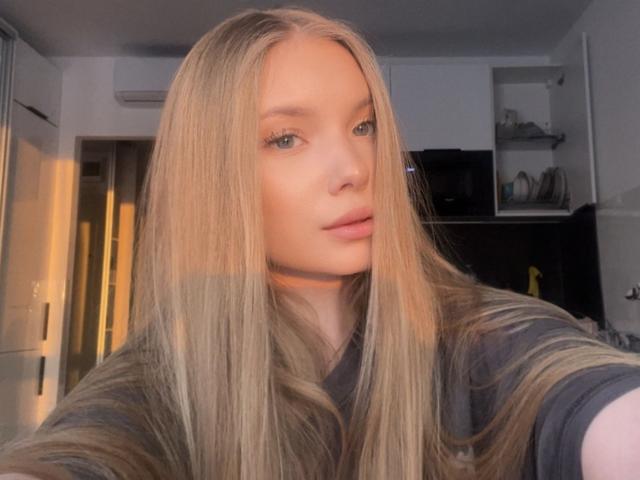 BarbraErrett has long, straight blonde hair, blue eyes, and pink lips. She's wearing a black shirt, with sunlight highlighting her face. Her pussy is not visible.