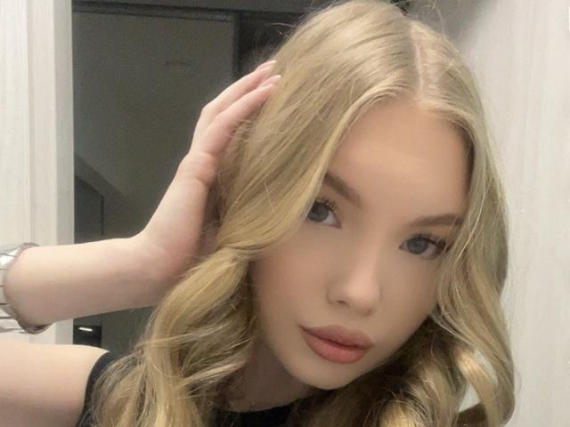 BarbraErrett with blonde wavy hair, light skin, and pink lips, looking at the camera, hand in hair, wearing a black top, wearing a silver wristwatch.