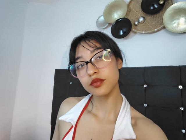 CamiVargas met een bril, rode lippenstift en een witte haltertop die haar borsten laat zien, zwart haar en Aziatische huid, staand voor een zwarte, gestoffeerde hoofdeinde..