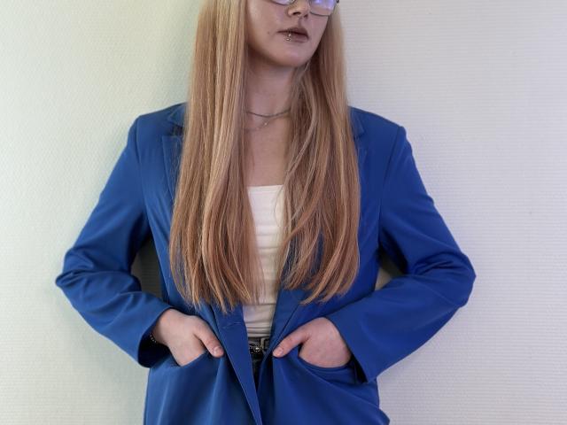 Piedad Mendenhall, con lunghi capelli biondi, indossa un blazer blu, una camicetta bianca, ha le mani nelle tasche, porta occhiali e una collana, ed è in piedi di fronte a un muro bianco..