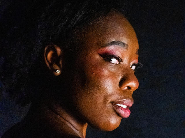 La piel marrón oscuro de JazaraKoral, la sombra de ojos roja y los labios rosados brillantes. Lleva pequeños pendientes, y tiene la cabeza ligeramente girada hacia un lado, mostrando su perfil..