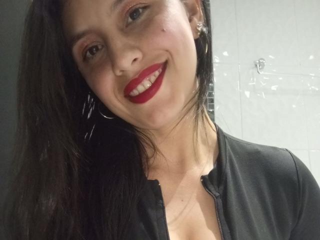 MadissonFontaine has dark brown hair, red lipstick, and is smiling at the camera with her black zip-up shirt showing a hint of cleavage. She's wearing large hoop earrings.