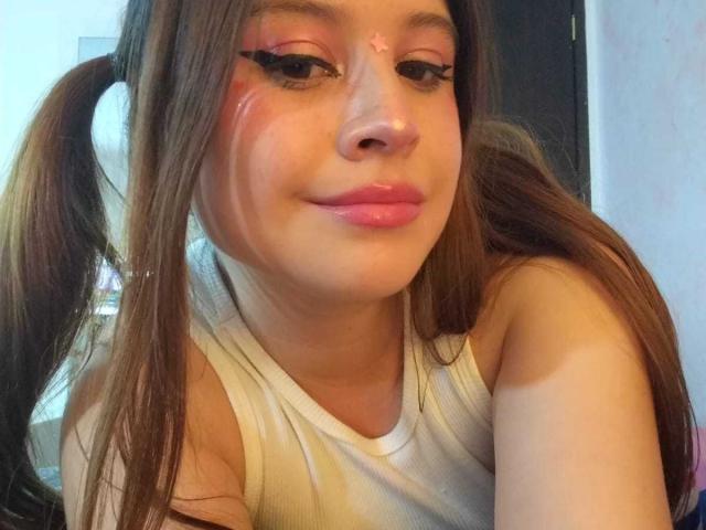 Melani Becker con eyeliner rosa, trucco occhi rosso e una stella sulla fronte, indossa una canottiera bianca, ha due trecce marroni, labbra rosa, sta sorridendo, in un selfie..