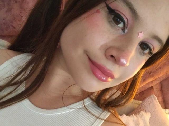 Le visage de Melani Becker, avec une étoile rose sur le front, portant un débardeur blanc, a des lèvres roses brillantes, de longs cheveux bruns et un eye-liner rose, et elle est allongée sur un tissu rose..