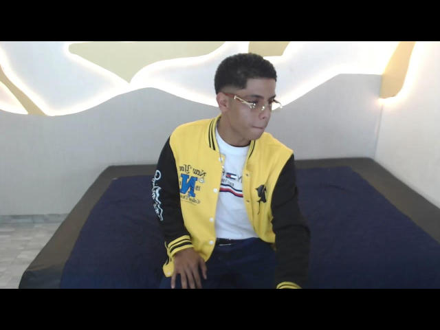 KingFastt with short black hair, glasses, yellow and black varsity jacket, white shirt, blue pants, sitting on dark bed, looking to the side, in a simple room.