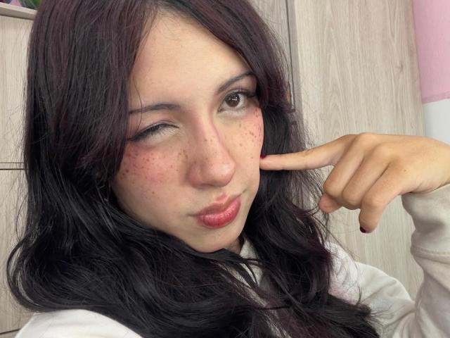 ValeryBeaumont with dark wavy hair, winking, and sticking her finger to her cheek, showing pink freckles and glossy red lips. She's wearing a white shirt.