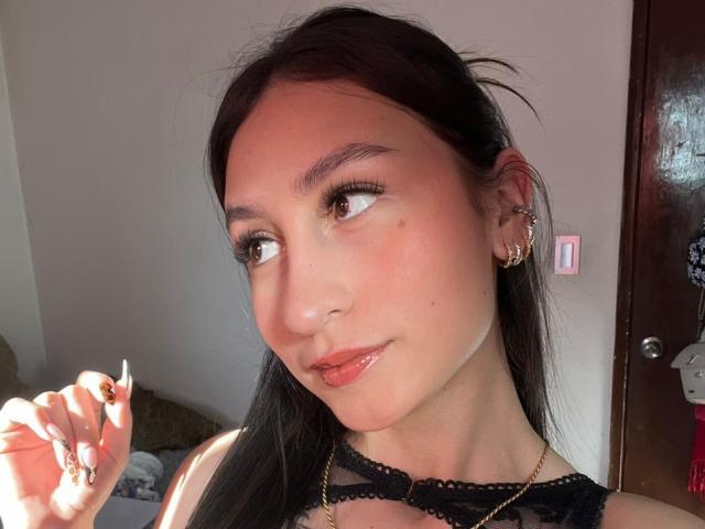 MaddyCarina with light brown skin, black hair, and subtle makeup, showing off her small nose and full pink lips, wearing black lace and gold earrings, holding a ring.
