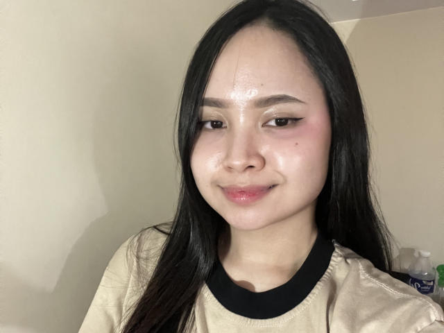 AlaineAtcher has shiny, sweaty skin on her face, black hair, and is wearing a beige shirt with a black collar, smiling slightly. Her brown eyes are half-open.