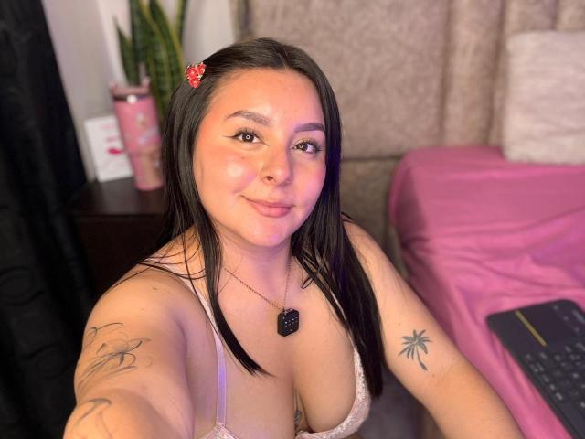 EmaJames, Latina with black hair, red hairclip, wearing lace bra, showing large tits, tattoos on arm, smiling, necklace with black gem, pink bed, laptop, potted plant.