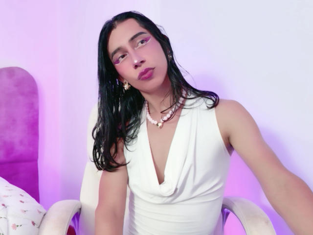 AlexaMissSx with long black hair, wearing a white sleeveless dress with a deep V-neck, pink eye makeup, and a pink necklace, sitting in a white chair.