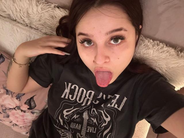 Lucie, met opvallend groene ogen en lang donkerbruin haar, met haar tong uitgestoken, draagt een zwart "Iron Maiden" shirt en kijkt omhoog naar de camera. Haar handen zitten op haar hoofd..