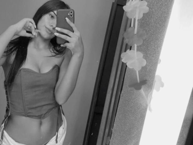 Autoportrait en noir et blanc d'AlaraKey avec de longs cheveux noirs, portant un haut sans bretelles qui dévoile ses seins et un léger ventre, mordant une ongle, tenant un téléphone..