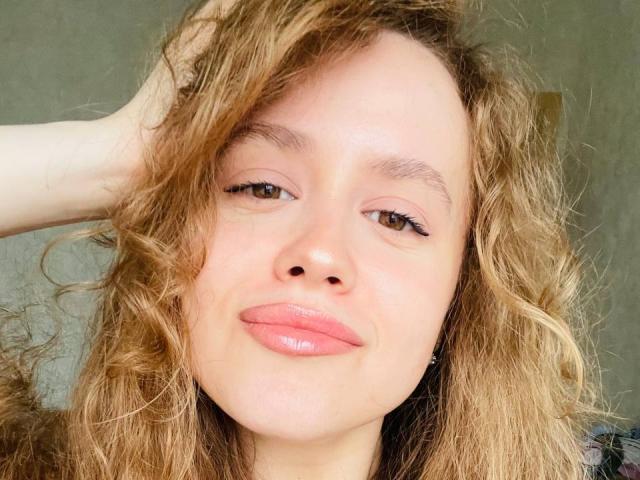 PhillisChinetti's face with curly blonde hair, pink glossy lips, and brown eyes, hand in hair, looking slightly up, showing a subtle smile. Her nose is small and cute.